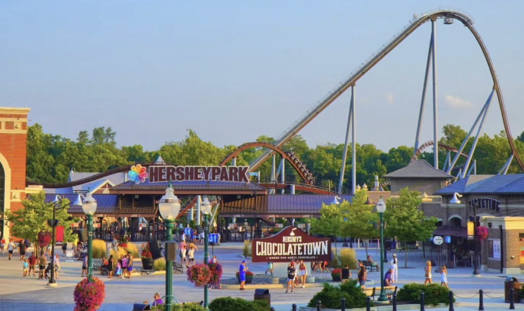 Entrance to Hersheypark on a sunny day, featuring the Hersheypark sign above the gates, the Chocolatetown welcome sign in the foreground, visitors walking through the plaza, and large roller coasters towering in the background. Hersheypark Fast Track Unlimited. 