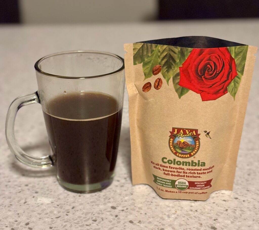 A cup of coffee next to a tan coffee pouch that says Java Planet Colombia