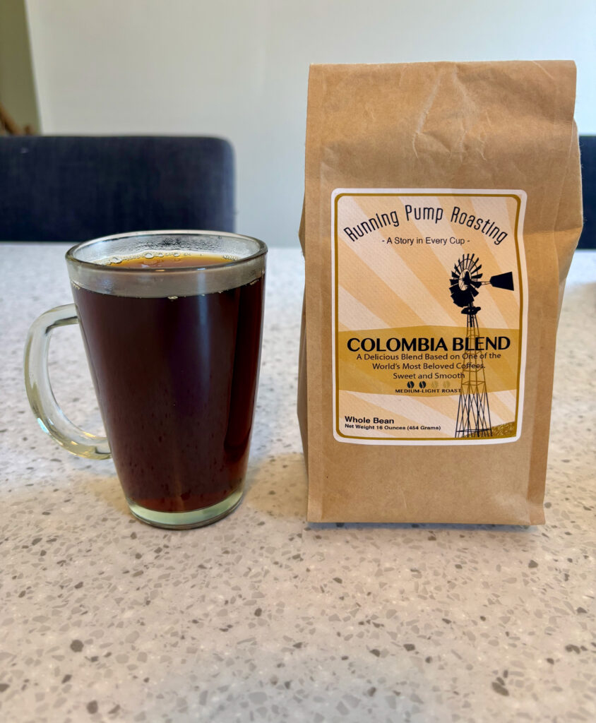 A clear glass mug filled with black coffee sits on a light speckled countertop next to a brown paper bag of “Running Pump Roasting Colombia Blend” whole bean coffee, labeled medium-light roast, 16 ounces.