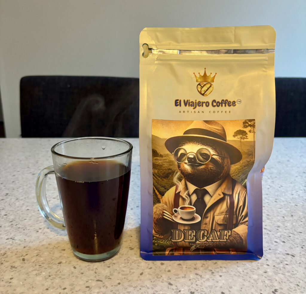 A clear glass mug filled with black coffee sits on a light countertop next to a beige-and-blue bag of El Viajero Coffee decaf, featuring an illustration of a sloth wearing glasses and a hat while holding a cup of coffee.
