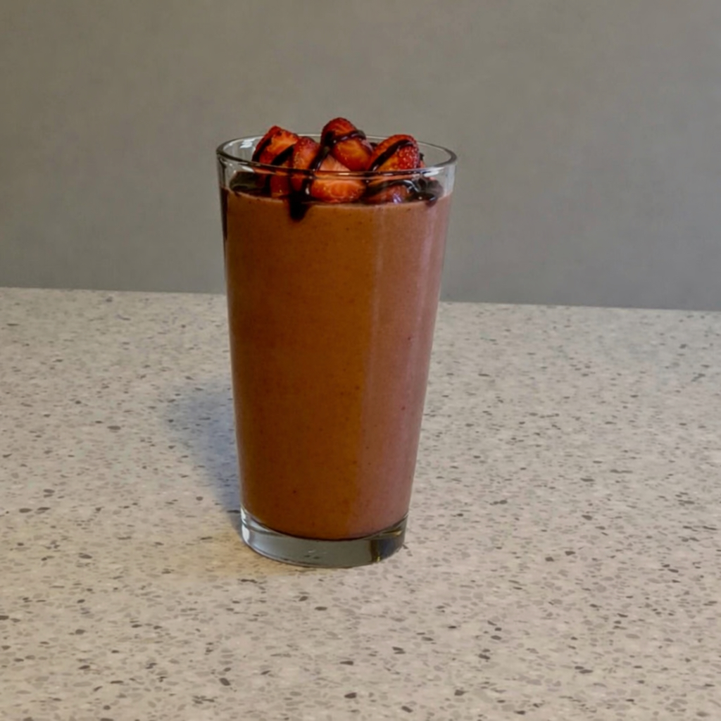 Chocolate Covered Strawberry Smoothie in a tall clear glass topped with fresh sliced strawberries and a drizzle of chocolate syrup, sitting on a light speckled countertop.