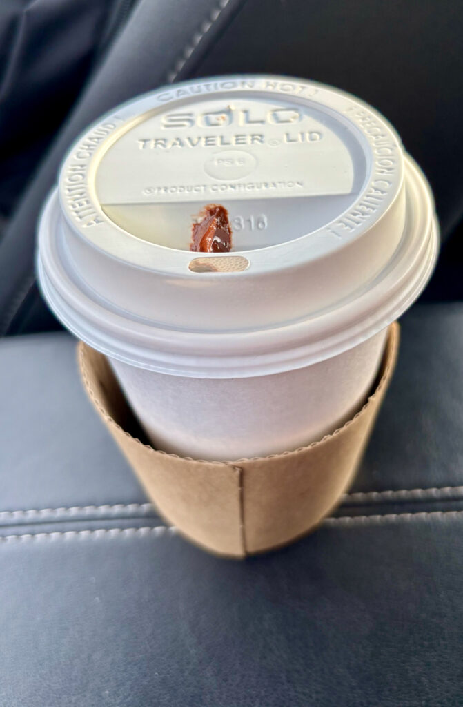 Close-up of a hot Americano in a white paper to-go cup with plastic lid and cardboard sleeve, resting inside a car.