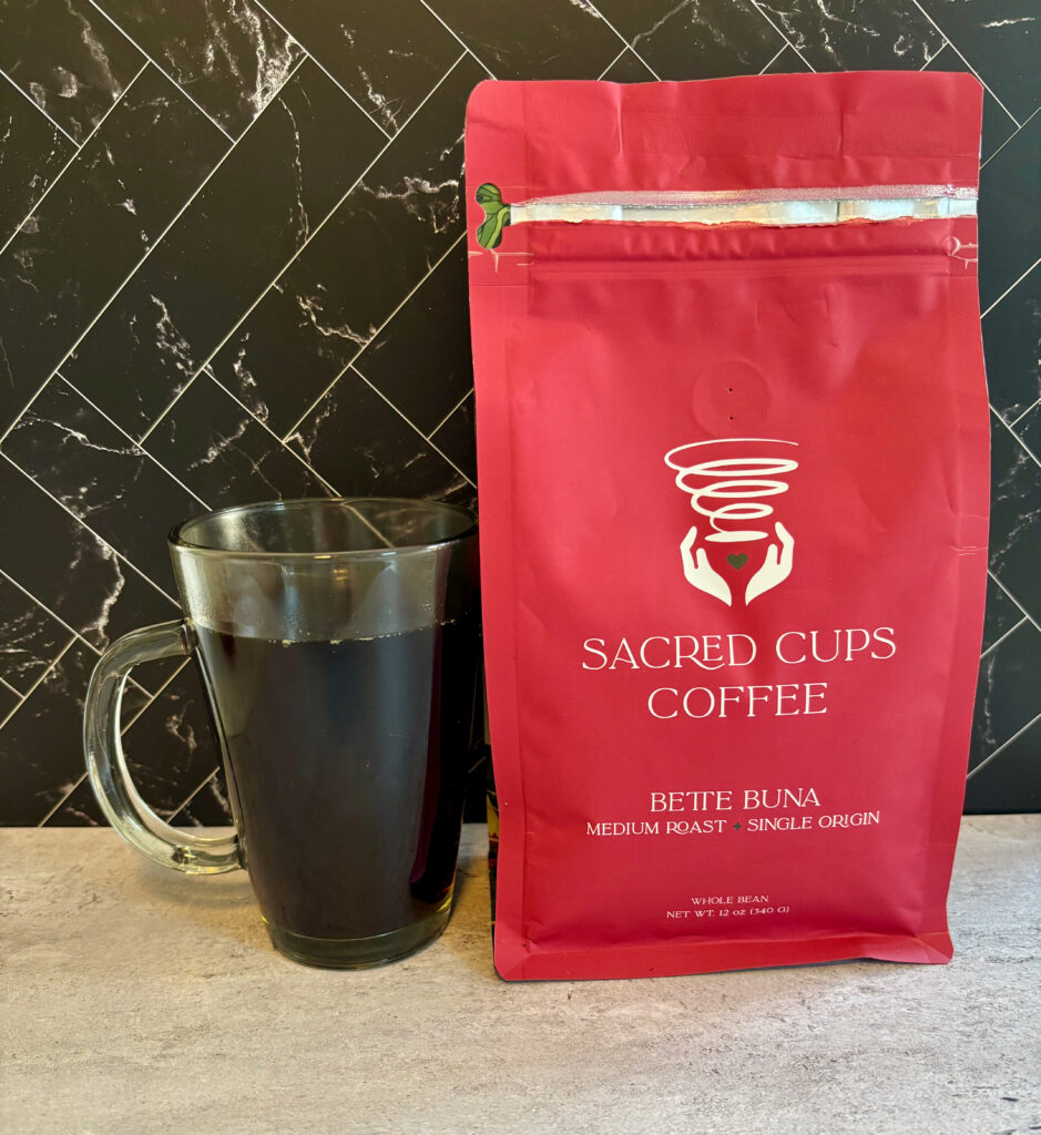 A cup of Sacred Cups Bette Buna coffee in a clear mug next to the bag of Bette Buna coffee. Poured for the Sacred Cups Bette Buna Review