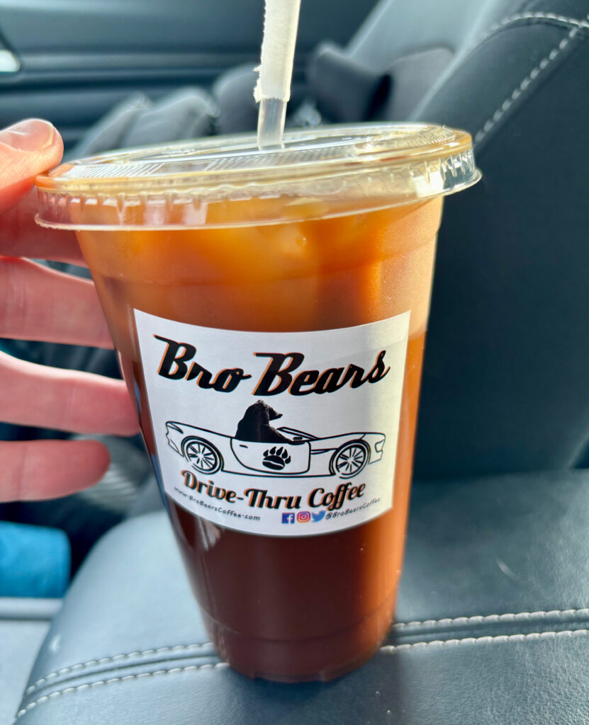 Iced Nutty Bro-Fessor coffee in a clear plastic cup with lid and straw, featuring the Bro Bears Drive-Thru Coffee logo, held inside a car.