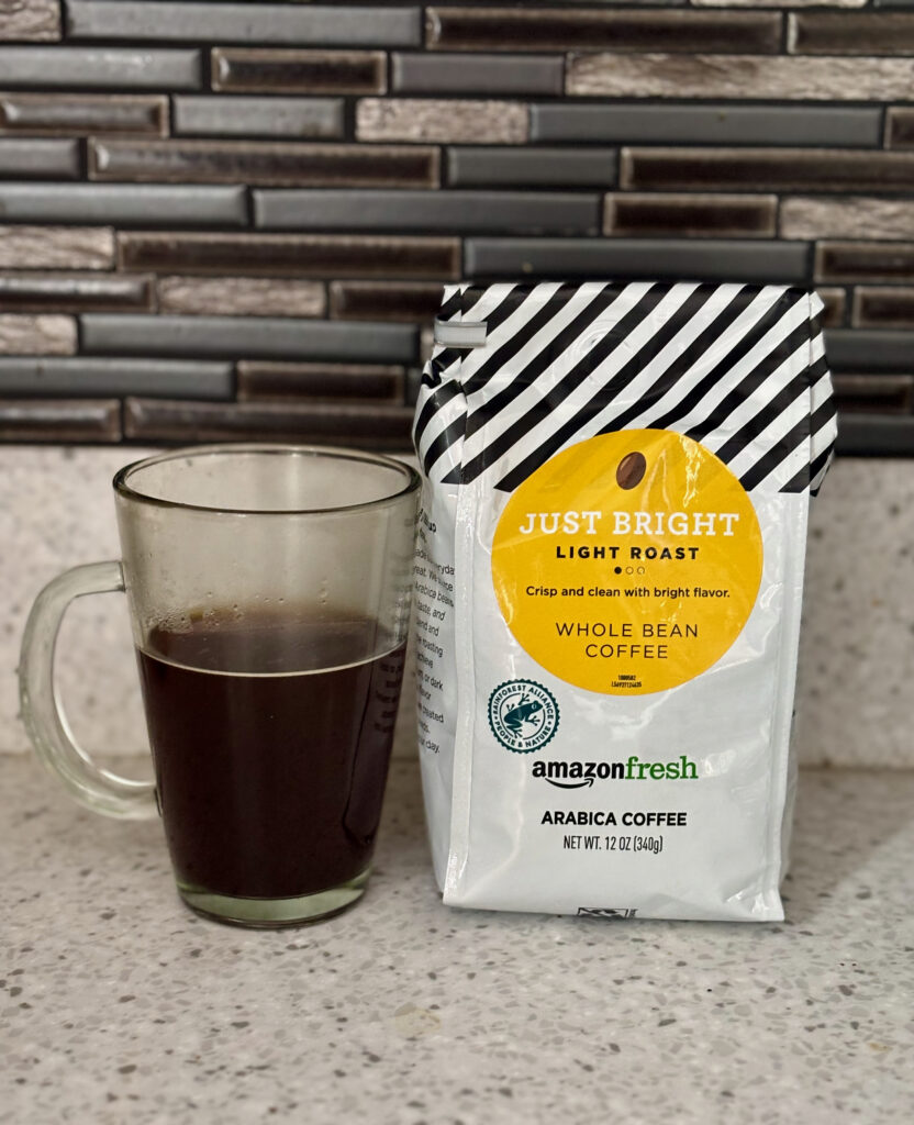 Amazon Fresh Just Bright Coffee Review photo showing a clear glass mug filled with brewed coffee next to an Amazon Fresh Just Bright coffee bag on a white countertop, with a black and gray thin tile backsplash in the background.