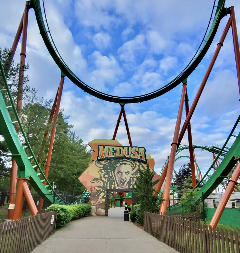 Entrance to the Medusa roller coaster, with green track curving overhead above the walkway and a large Medusa-themed sign beneath a cloudy blue sky for a Six Flags Great Adventure review