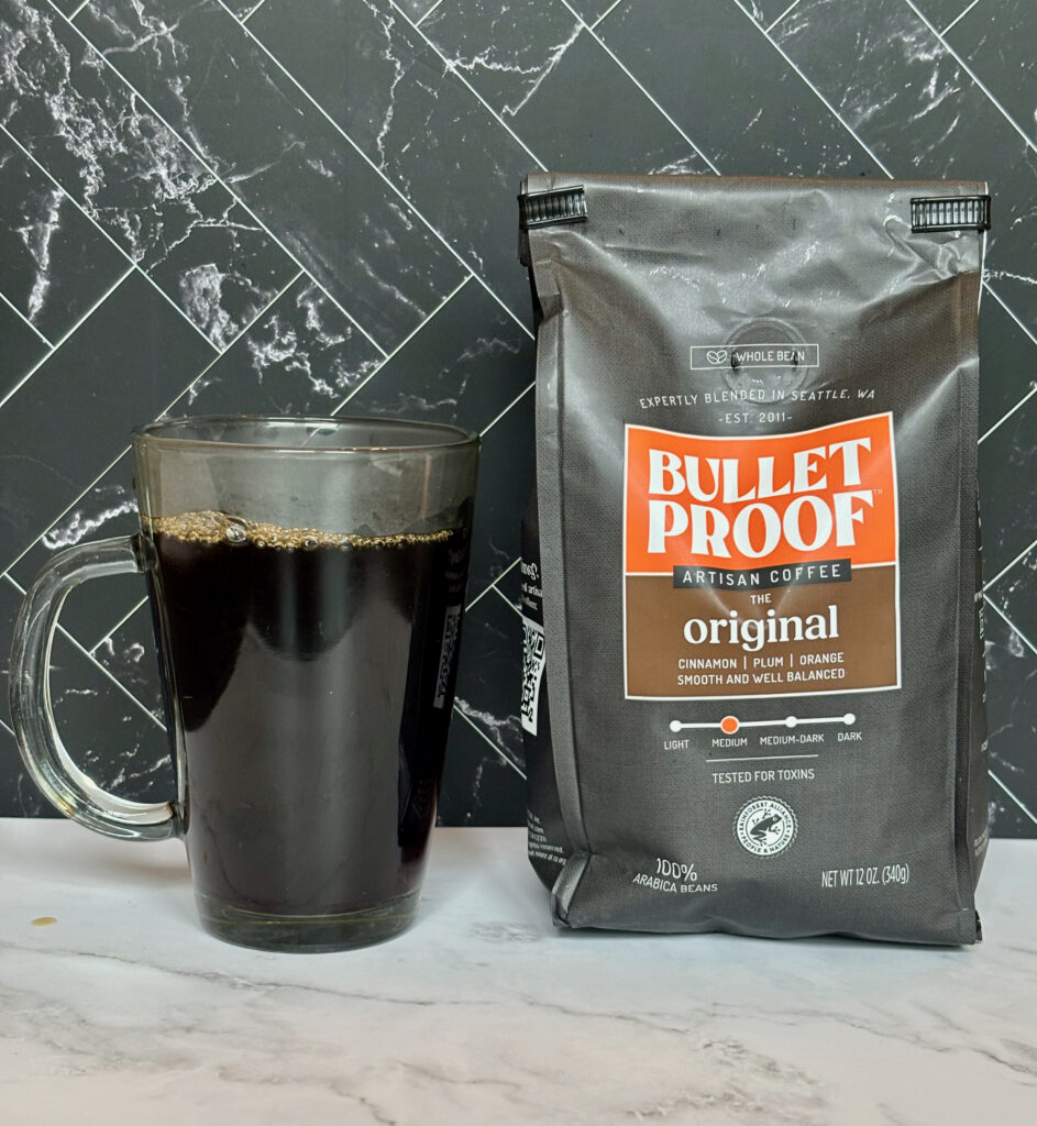A cup of black coffee for Bulletproof Coffee The Original Review sitting in a clear mug next to the black bag 