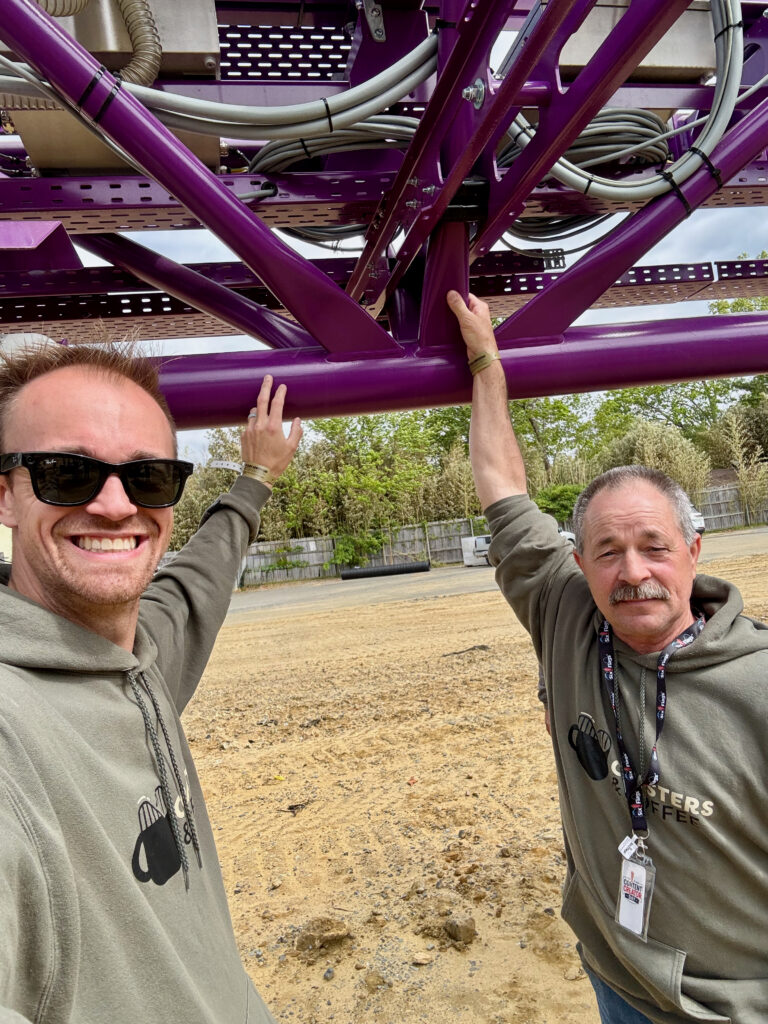 Two men stand beneath a purple roller coaster track under construction, each reaching up to touch the support structure during a close-up visit to the future ride site for a Six Flags Great Adventure review.