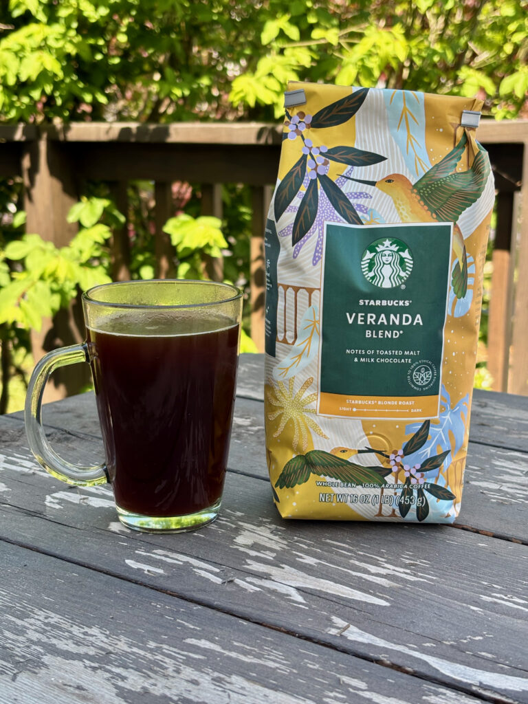 Starbucks Veranda Blend Coffee Review photo showing a cup of Starbucks Veranda poured into a clear mug sitting next to the bag of Starbucks Veranda. Both sit on a wooden table on a deck