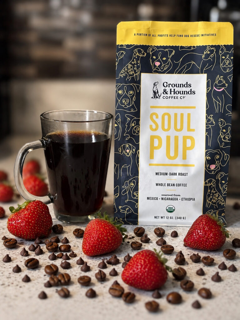 A bag of Grounds & Hounds Soul Pup coffee beside a glass mug of black coffee, surrounded by fresh strawberries, coffee beans, and chocolate chips on a kitchen countertop in this Grounds & Hounds Soul Pup review.