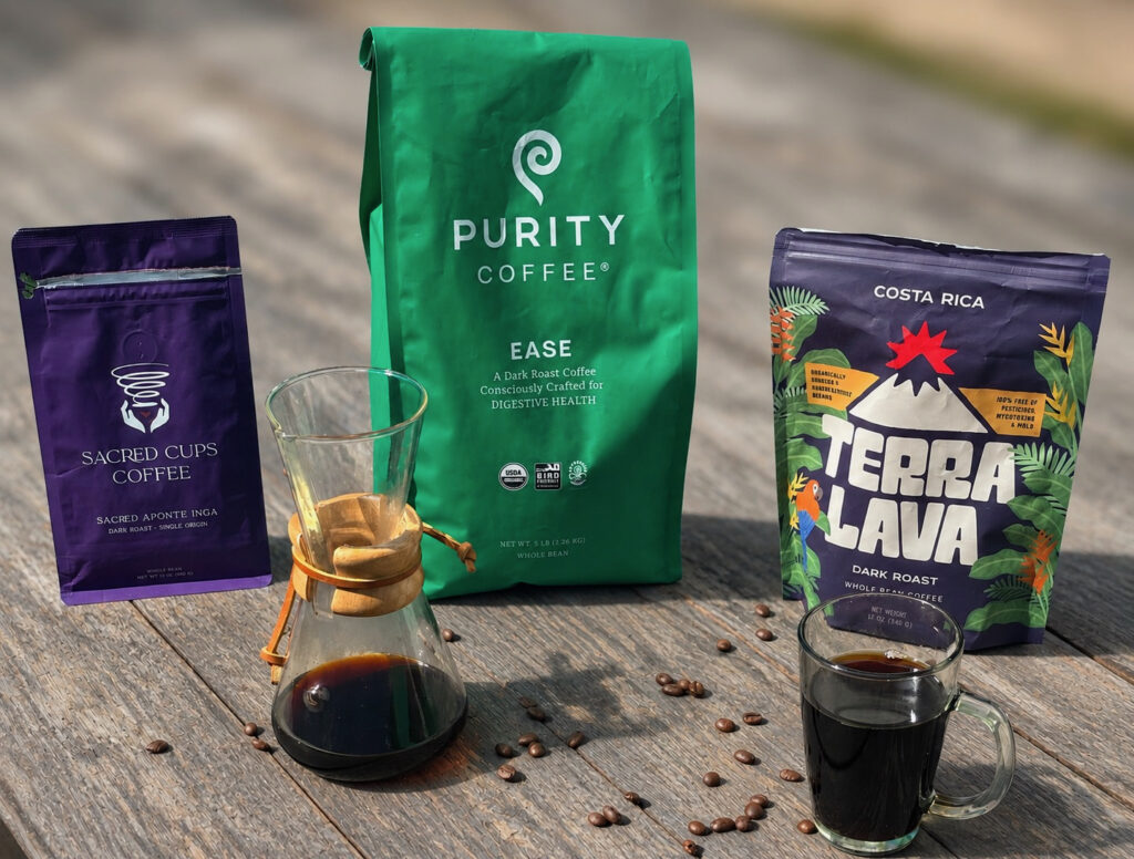 Alt Text:
5 Best Dark Roast Coffees of 2026 comparison featuring three premium dark roast coffee bags arranged on a rustic wooden table with scattered coffee beans, a glass pour-over coffee maker filled with freshly brewed coffee, and a clear glass mug of black coffee. The background is softly blurred to keep the focus on the coffee products and brewing setup in bright natural sunlight.