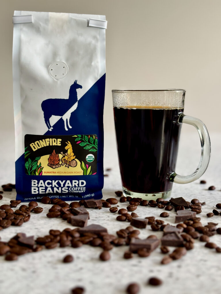 Backyard Beans Bonfire Coffee Review featuring a bag of Bonfire Sumatra medium-dark roast coffee beside a glass mug of black coffee with roasted coffee beans and dark chocolate scattered across the countertop.