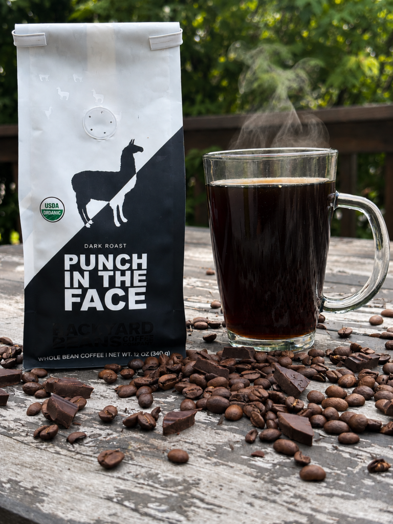 Backyard Beans Punch in the Face Review image showing a bag of Backyard Beans Punch in the Face dark roast beside a steaming glass mug of black coffee with coffee beans and dark chocolate pieces on a rustic outdoor table.